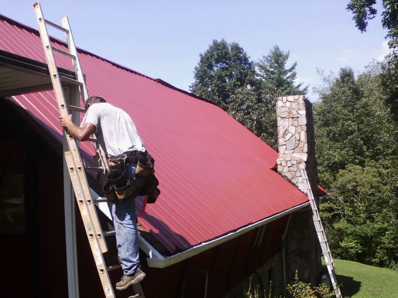 Roofer on a ladder beside a red metal roof and stone chimney — Soffit & Fascia Repair by Roofing Brothers in Rives Junction